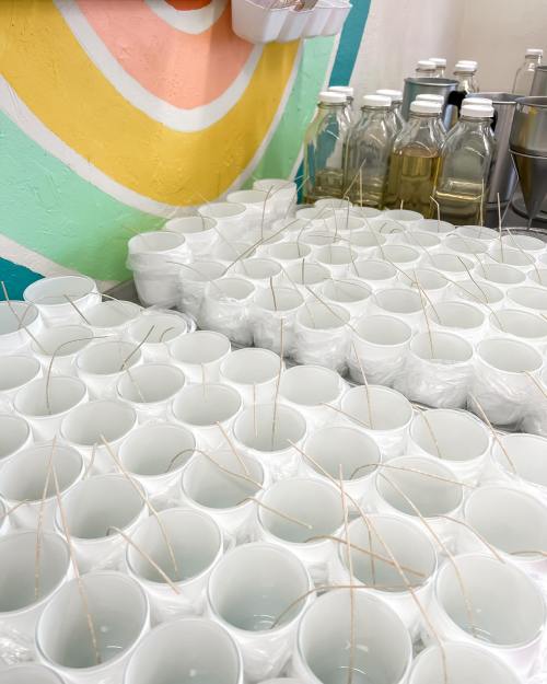 A table filled with white candle cups, each with a wooden wick, beside glass bottles of liquid. Colorful wall in background.