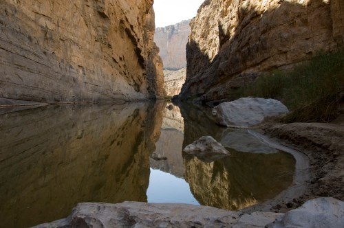 A serene river flows between towering rock formations, reflecting the cliffs in calm water.