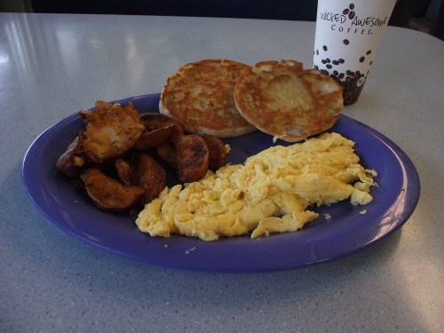 A plate with scrambled eggs, seasoned potato wedges, and two toasted English muffins, alongside a coffee cup.
