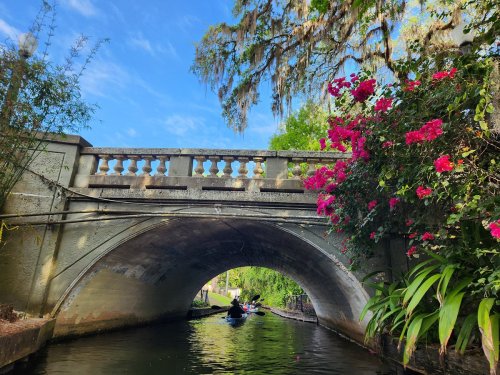 A serene canal scene with a stone bridge, vibrant flowers, and a person kayaking under a blue sky.