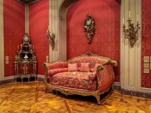 Luxurious pink sofa with ornate gold detailing, set against a rich red wall, accompanied by a decorative clock and wall sconces.