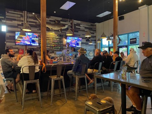 A lively bar scene with patrons seated at the counter, watching TVs and enjoying drinks in a casual atmosphere.