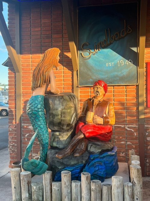 A colorful mermaid statue interacts with a seated man in a red turban outside a shop named "Sinbad's."