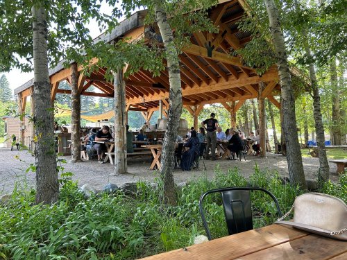 A rustic outdoor dining area with wooden structures, surrounded by trees and people enjoying their meals.
