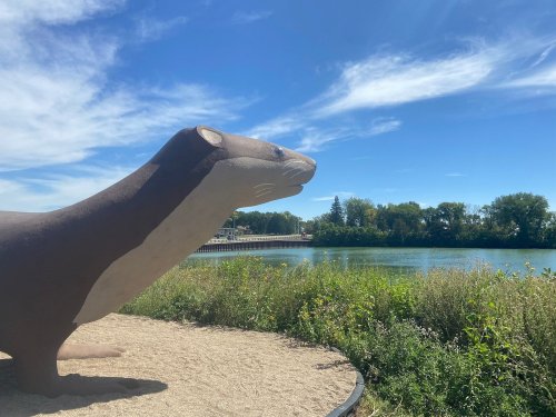 A large otter sculpture overlooks a river with blue skies and greenery in the background.