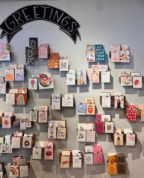 A wall display of colorful greeting cards with various designs and messages, topped with a "Greetings" sign.