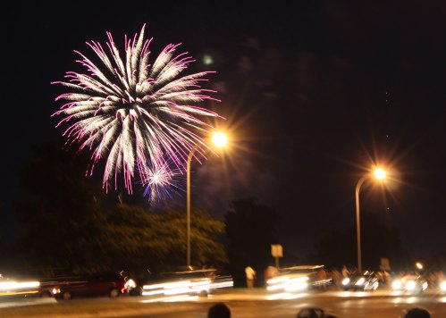 Colorful fireworks burst in the night sky above a busy street, with car lights streaking by below.
