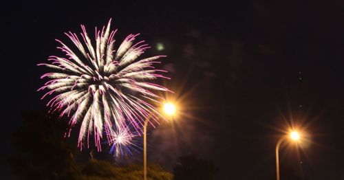 Colorful fireworks burst in the night sky, illuminating the scene with bright pink and white sparks. Streetlights glow below.