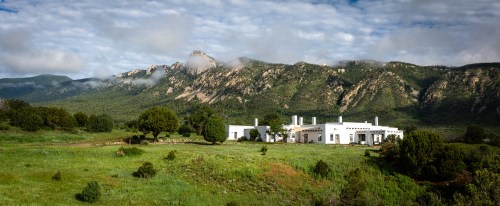 A white building surrounded by green fields, with mountains and clouds in the background.
