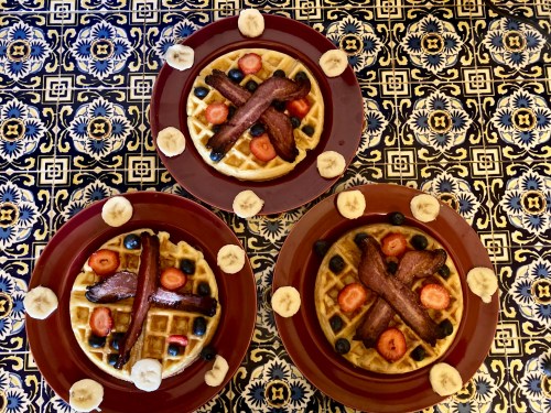 Three plates of waffles topped with bacon, strawberries, blueberries, and banana slices, on a patterned tablecloth.