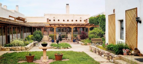 A serene courtyard with stone pathways, greenery, and a central fountain, surrounded by adobe-style buildings.