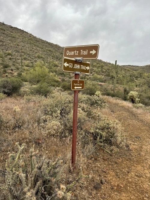 Trail sign indicating directions for Quartz Trail and Go John Trail in a desert landscape.