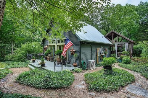 A charming gray cottage surrounded by lush greenery, featuring a porch, American flag, and decorative plants.