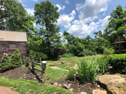 A scenic garden with lush greenery, a wooden shed, and a blue sky dotted with clouds.
