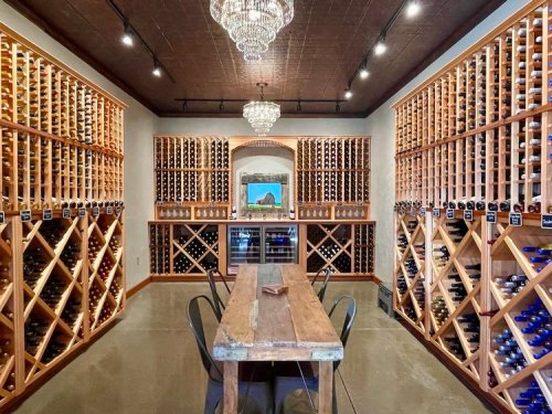 A wine cellar featuring wooden racks filled with bottles, a central table, and a chandelier overhead.