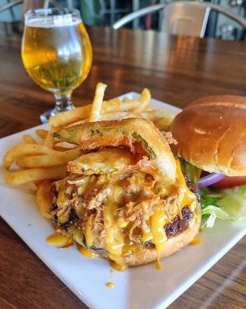 A plate with a cheesy burger topped with onion rings, fries, and a glass of beer in the background.