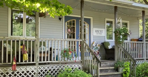 A charming cottage with a welcoming porch, decorated with plants and cozy seating, surrounded by greenery.