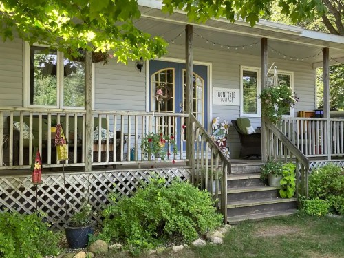 A cozy porch with plants, seating, and decorative elements, surrounded by greenery and sunlight.