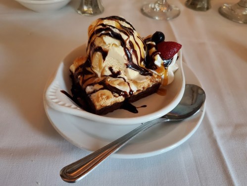 A dessert bowl with a brownie topped with vanilla ice cream, chocolate syrup, and a strawberry, served with a spoon.