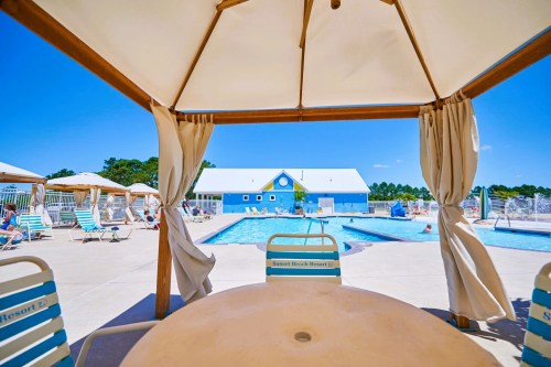 View of a sunny pool area with lounge chairs, cabanas, and a blue building in the background.