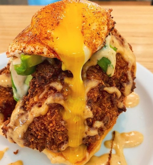 A close-up of a fried chicken sandwich topped with a runny egg, jalapeños, and drizzled with sauce on a white plate.