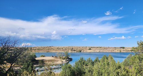 A serene lake surrounded by greenery and hills under a blue sky with scattered clouds.
