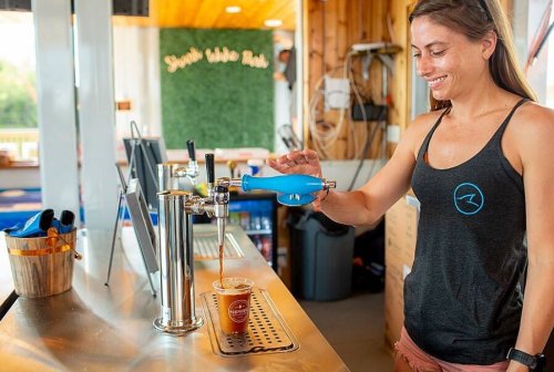 A woman in a tank top pours a drink from a tap into a cup at a bar.