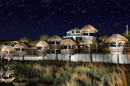 A waterfront restaurant with thatched roofs under a starry night sky, reflecting in the calm water below.