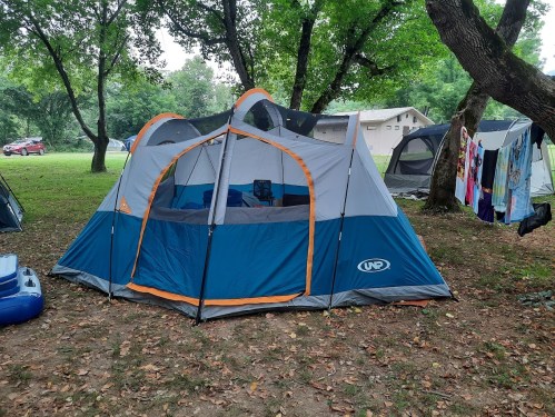 A large blue and gray tent set up in a grassy area, surrounded by trees and other camping gear.