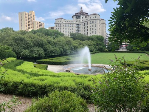 A fountain sprays water in a landscaped park, surrounded by lush greenery and a historic building in the background.