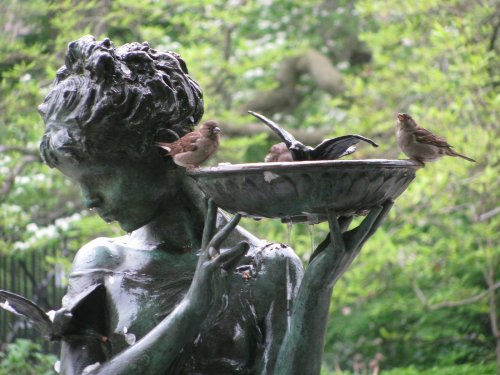 A bronze statue of a woman holding a birdbath, with several birds perched on the edge and around her.