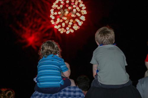 Two children sit on a person's shoulders, watching vibrant red fireworks against a dark night sky.