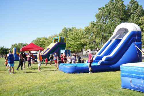 A sunny park scene with people enjoying inflatable water slides and a bounce house. Trees in the background.