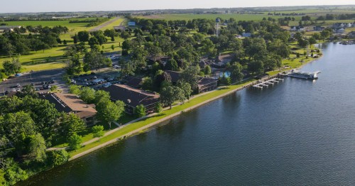 Aerial view of a lakeside community with green trees, buildings, and a parking area along the water's edge.