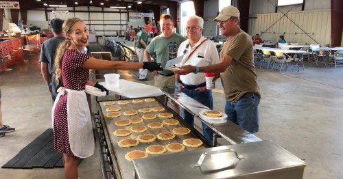 A young woman in an apron serves pancakes to three smiling men at a community event in a large indoor space.