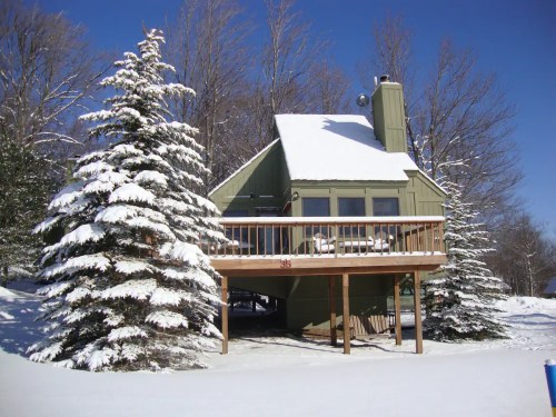A cozy green cabin surrounded by snow-covered trees under a clear blue sky.