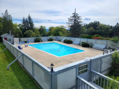 A clear blue swimming pool surrounded by a fenced area, with lounge chairs and greenery in the background.
