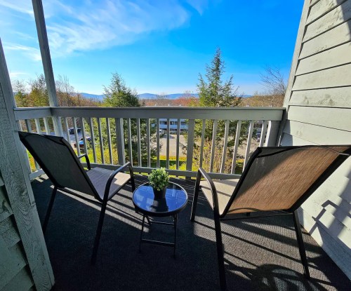 A balcony with two chairs and a small table, overlooking a scenic view of trees and mountains under a clear blue sky.