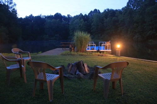 A serene lakeside scene at dusk, featuring a dock, chairs around a fire pit, and lush greenery.