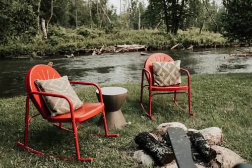 Two red chairs with pillows sit by a river, next to a fire pit surrounded by rocks and greenery.