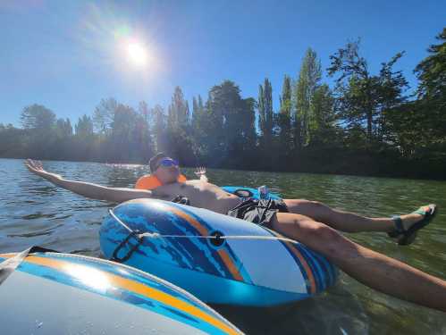 A person relaxes on a float in a river, arms outstretched, under a bright sun with trees in the background.