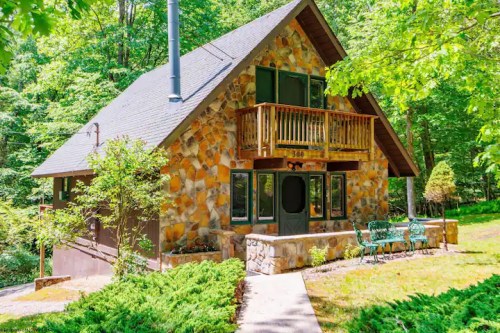 A charming stone cottage surrounded by lush greenery, featuring a balcony and a welcoming front path.