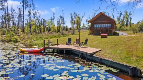 A serene lakeside scene featuring a wooden cabin, dock, kayak, and chairs surrounded by lily pads and trees.