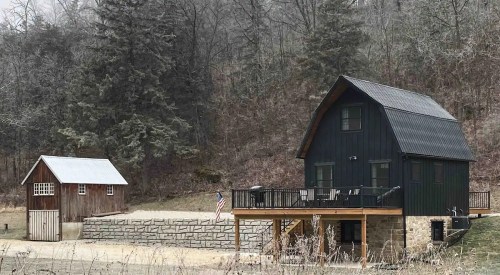 A modern black house with a wooden deck next to a rustic barn, surrounded by trees and a gravel area.