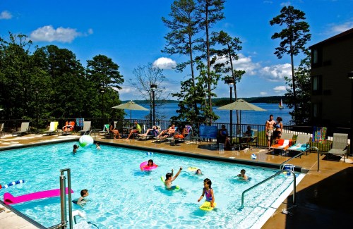 A sunny pool scene with people swimming and relaxing by the water, surrounded by trees and a lake in the background.