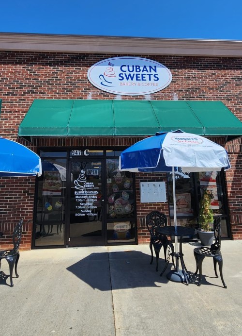 Exterior of Cuban Sweets bakery with a green awning, outdoor seating, and a sign displaying hours and menu.