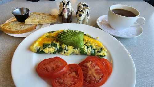 A plate with a spinach and cheese omelette, sliced tomatoes, toast, a cup of coffee, and two decorative figurines.