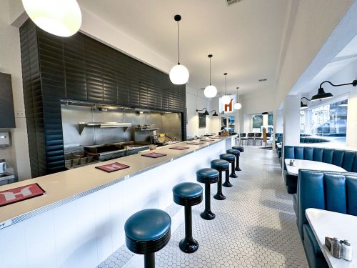 A modern diner interior featuring a long counter with black stools, white walls, and pendant lighting.