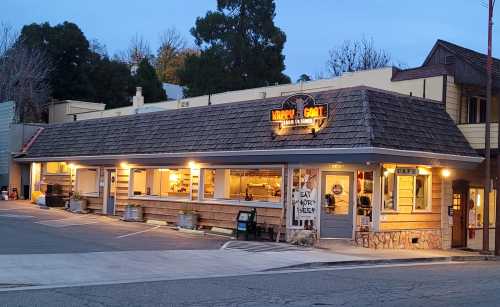 A cozy café with a shingled roof, warm lights, and outdoor seating, set in a quiet street at dusk.