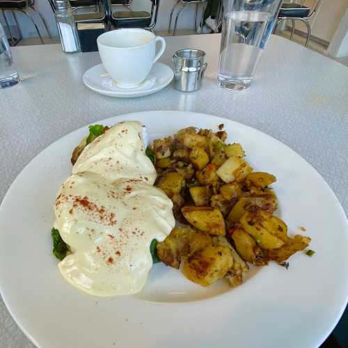 A plate of eggs Benedict with creamy sauce, served with seasoned roasted potatoes, alongside a cup of coffee and water.